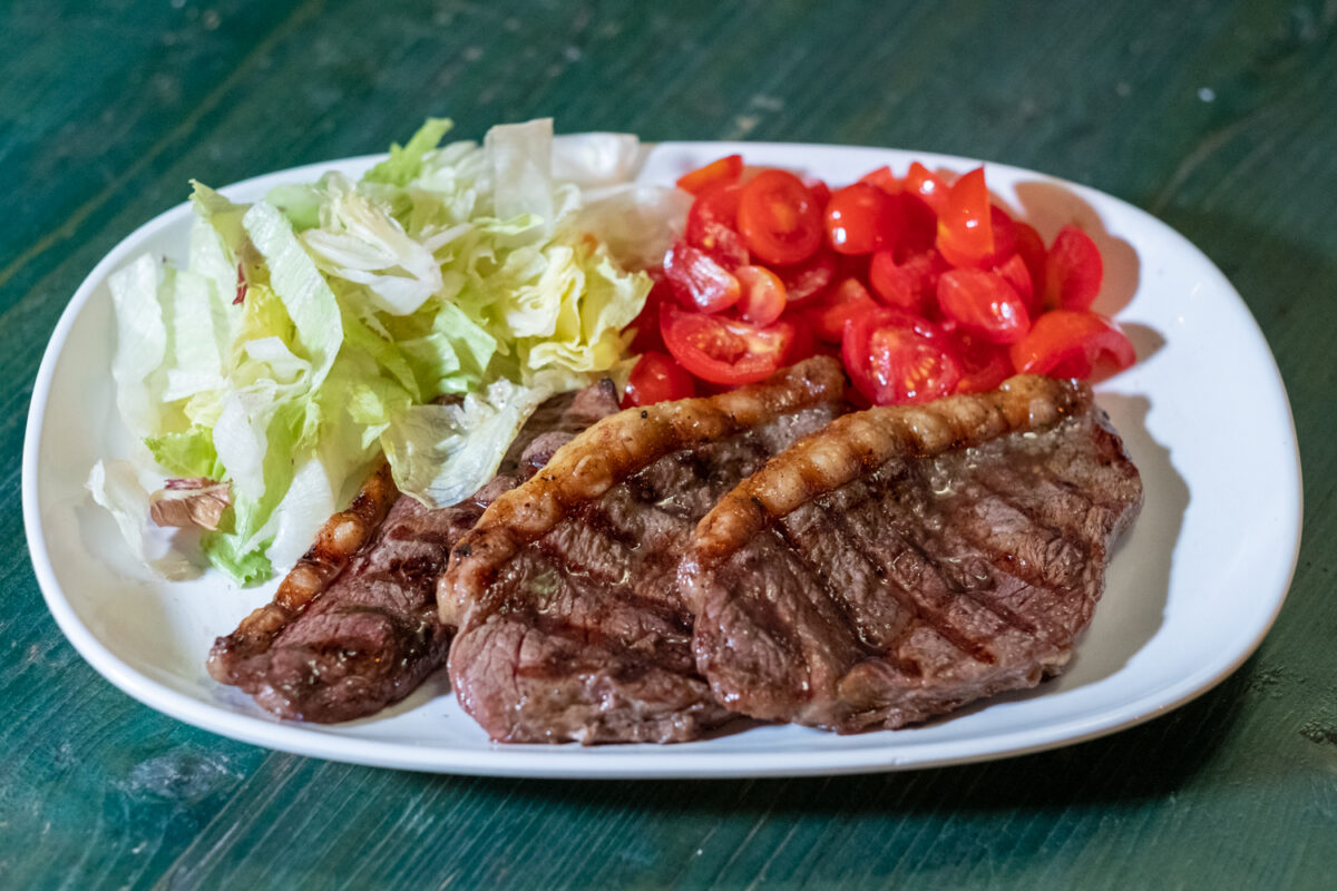Tre fette di bistecca alla griglia con insalata e pomodorini a spicchi, su un piatto ovale bianco