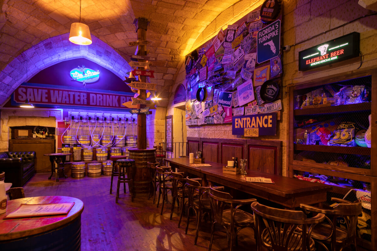 La cantina del Road 66: fusti d'acciaio allineati sotto la volta in pietra, l'insegna al neon Save Water Drink Beer e il muro coperto di targhe automobilistiche americane
