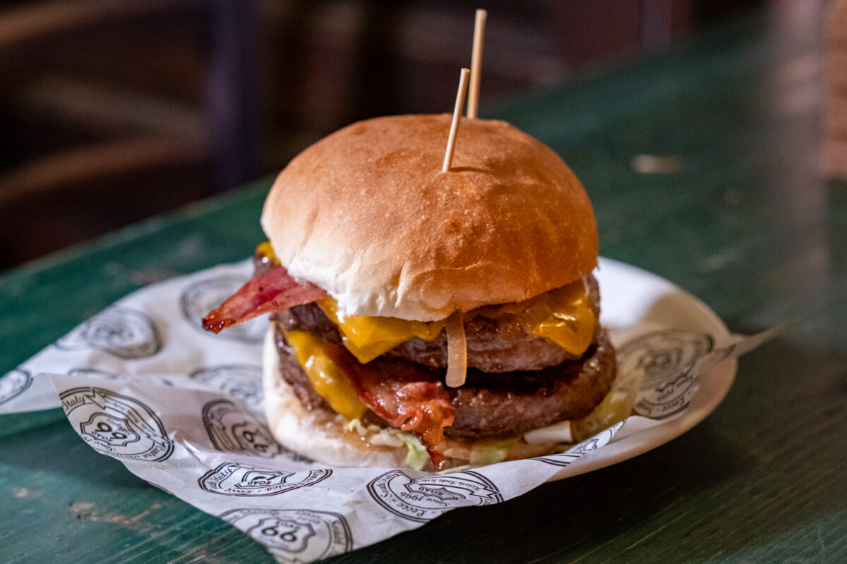 Hamburger doppio con bacon croccante e cheddar fuso, servito su carta oleata stampata con lo scudo Road 66