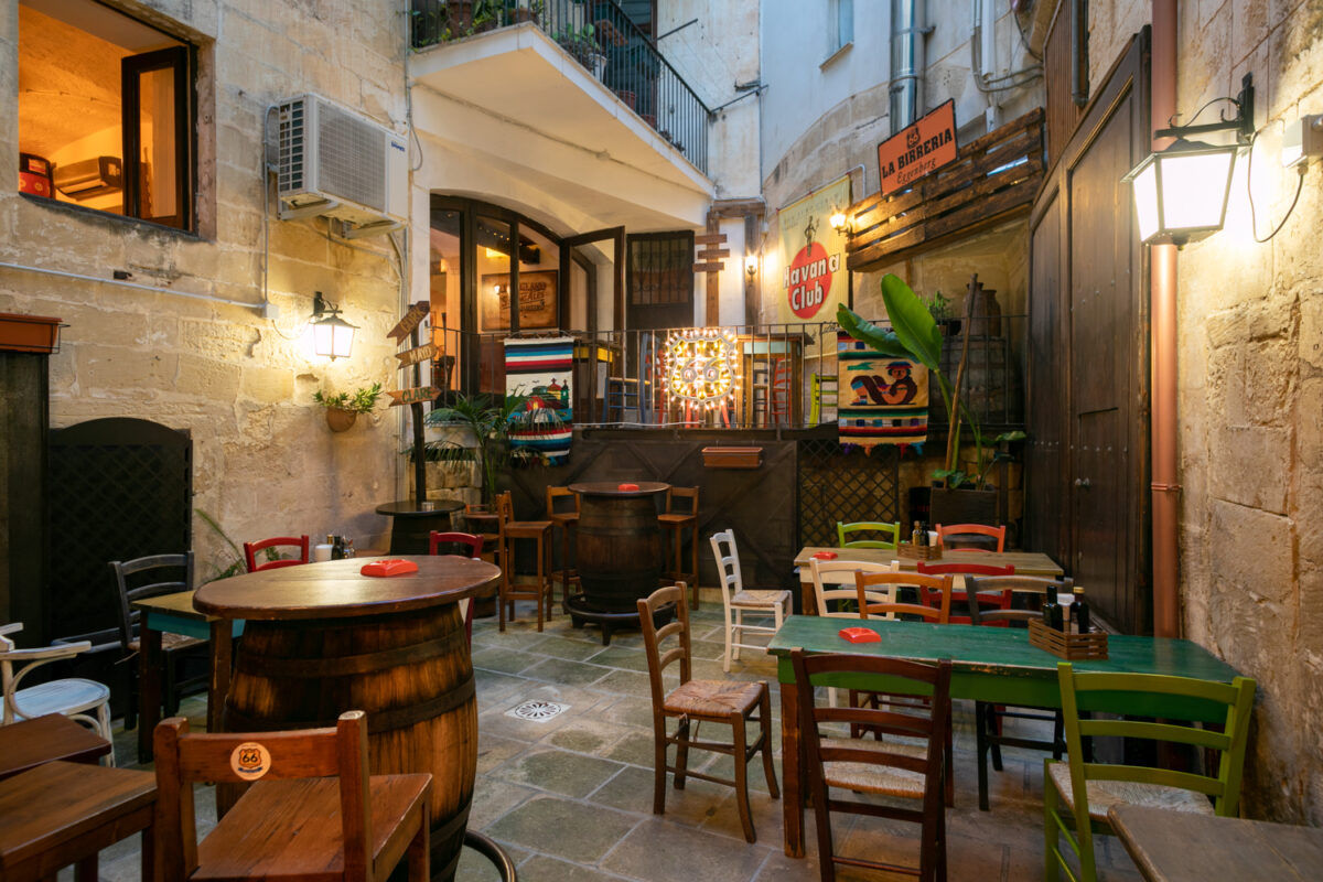 Il cortile interno del Road 66 con tavoli su botti, sedie colorate, l'insegna La Birreria Eggenberg e le pareti in pietra leccese