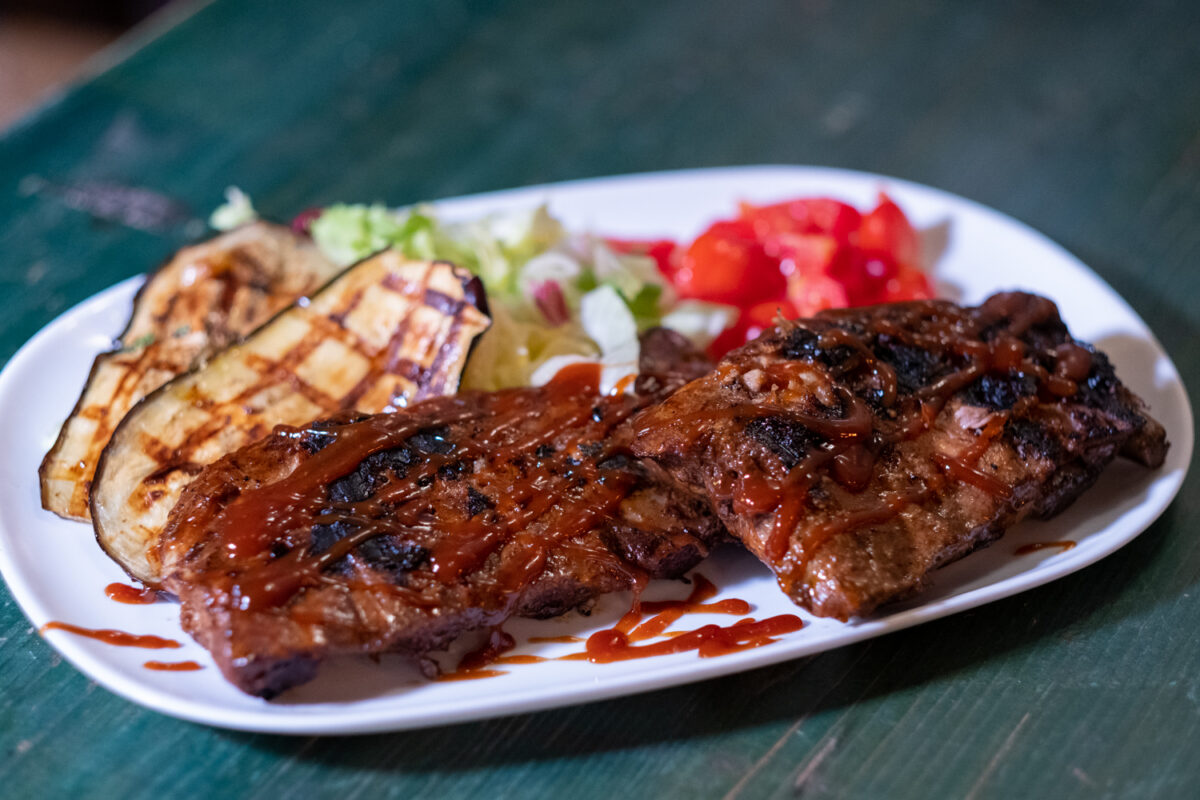 Costine di maiale glassate con salsa barbecue, servite con melanzana grigliata, insalata e pomodoro su un piatto bianco