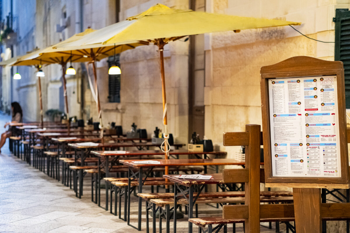 I tavoli da birreria del Road 66 in fila sotto gli ombrelloni gialli lungo la parete in pietra, con la bacheca del menu in legno