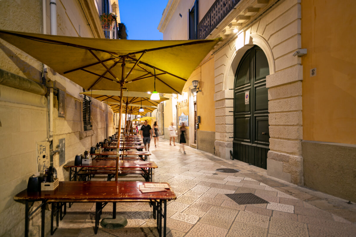 Via dei Perroni al tramonto con gli ombrelloni gialli del Road 66, i tavoli da birreria lungo il muro in pietra e il portone verde del civico 8