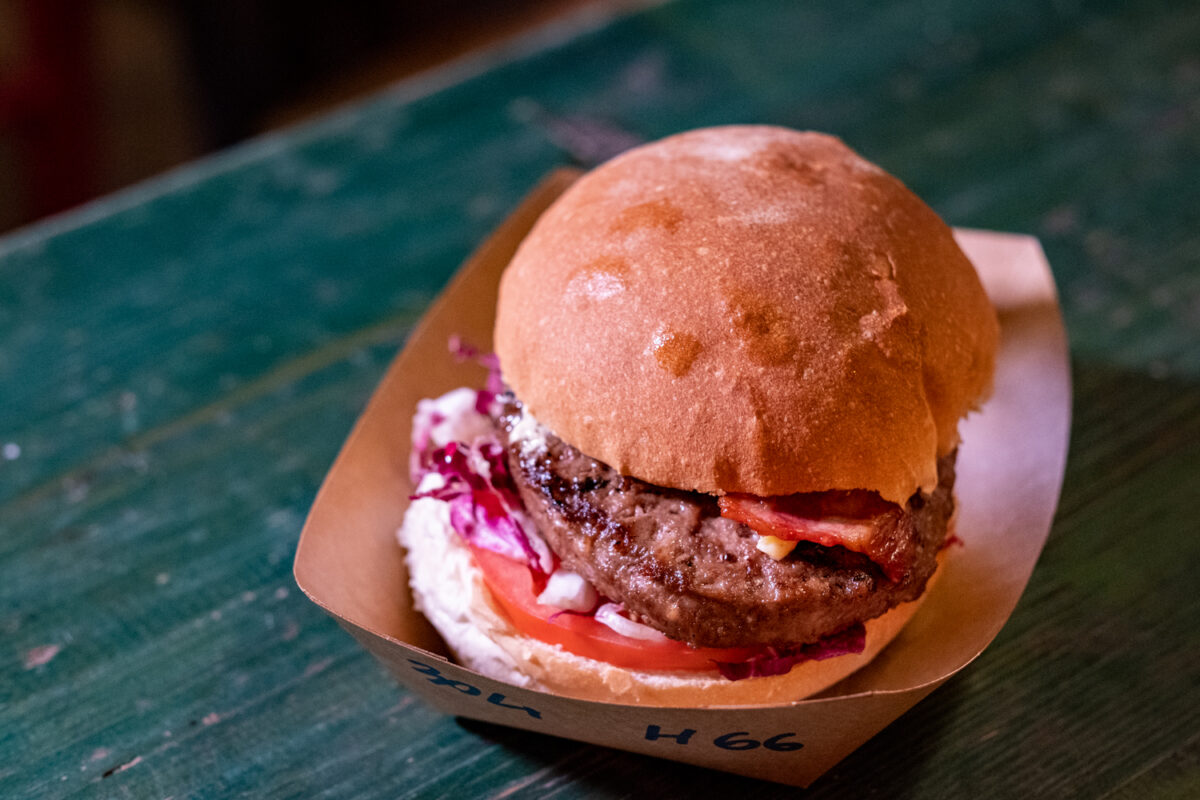 Hamburger con bacon, pomodoro e radicchio servito in un vassoietto di carta marcato 66, su un tavolo di legno verde del Road 66