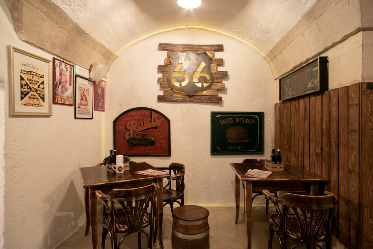 Sala a volta bianca del Road 66 con lo scudo 66 in metallo retroilluminato al centro della parete, insegne vintage Buick e Whisky e tavolini di legno