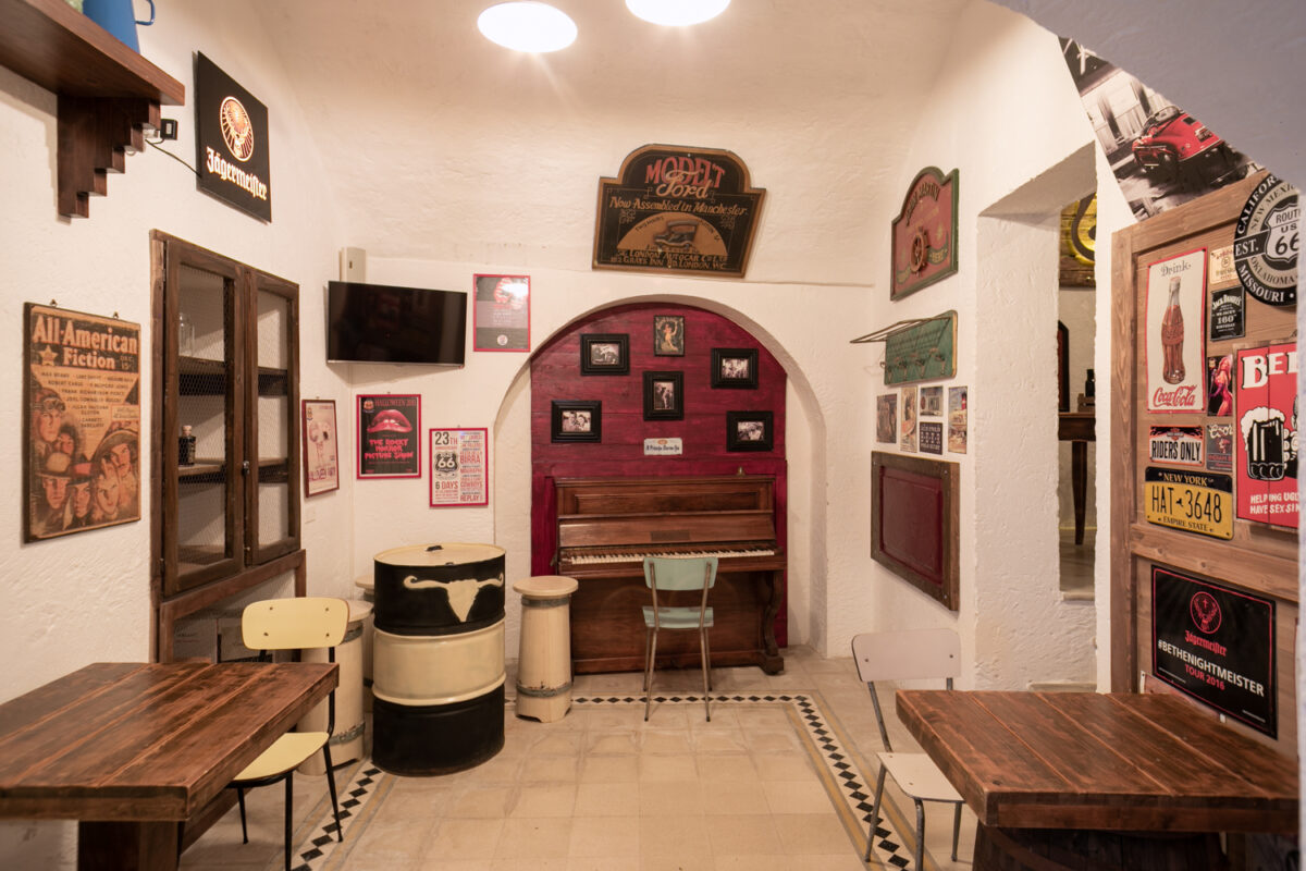 Sala del Road 66 con un pianoforte verticale dentro una nicchia rossa, manifesti americani e un bidone decorato con un teschio di bovino